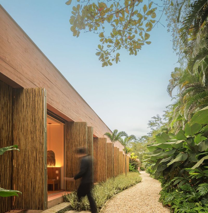 Bamboo-clad facade with illuminated doorway and figure walking along gravel path at dusk
