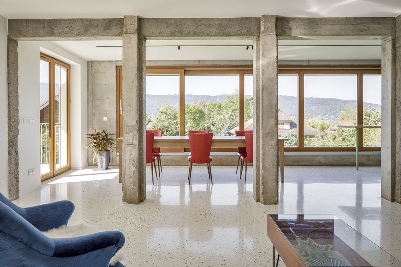 Through the columns: concrete structure framing the dining area with red chairs, full-height timber windows, mountain view