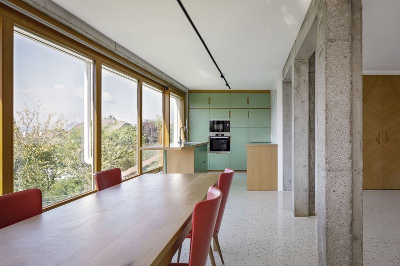 Kitchen and dining: sage green cabinets, long timber table with red chairs, concrete column, mountain view through windows