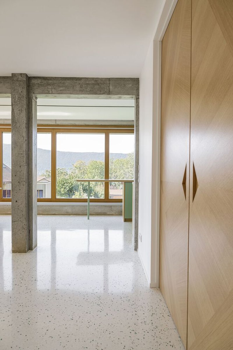 Corridor: timber wardrobe, concrete beam overhead, terrazzo floor, green kitchen glimpsed through the opening