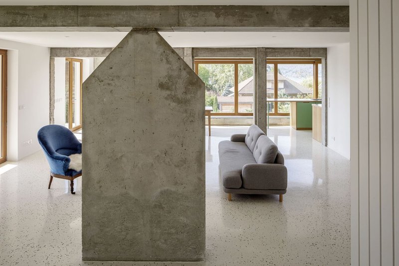 Living room from fireplace: house-shaped concrete chimney, grey sofa behind, timber doors, mountain light