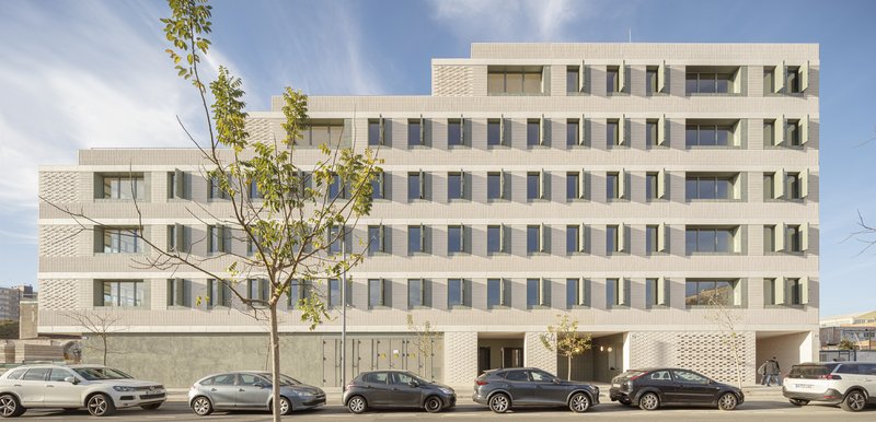 Street facade: six-storey cream brick building with stepped upper levels, perforated ceramic screen at ground floor, parked cars, young tree