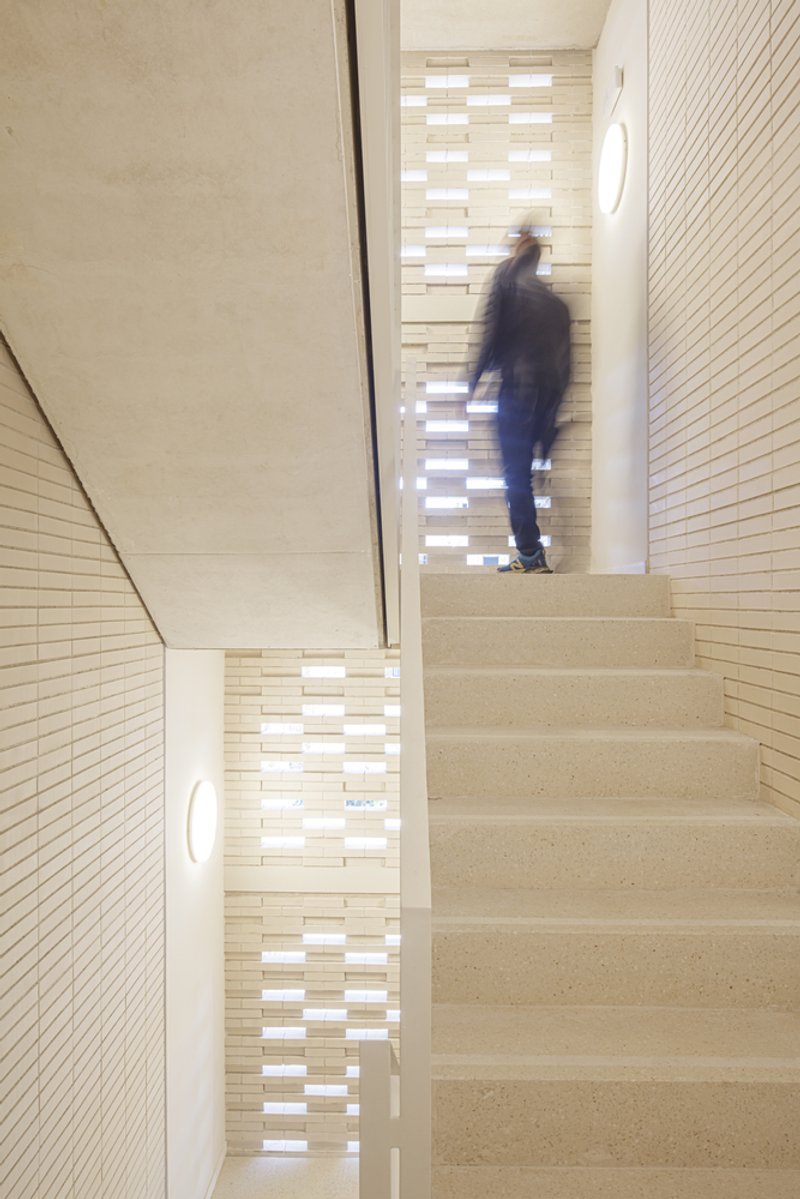Stairwell: figure ascending concrete stairs, perforated ceramic screen casting patterned light on the walls, timber handrail