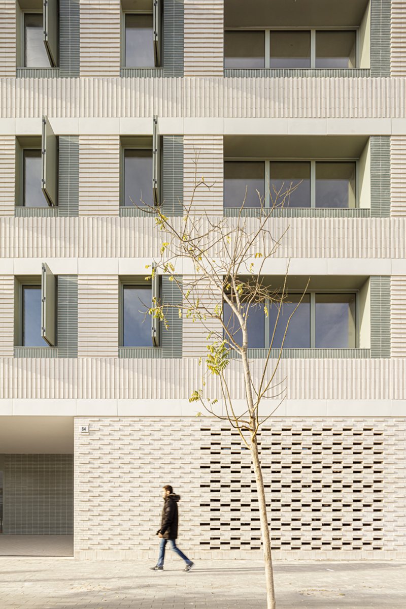 Street facade from the west: cream brick with perforated ceramic base, regular window grid, figure walking, young tree on the pavement
