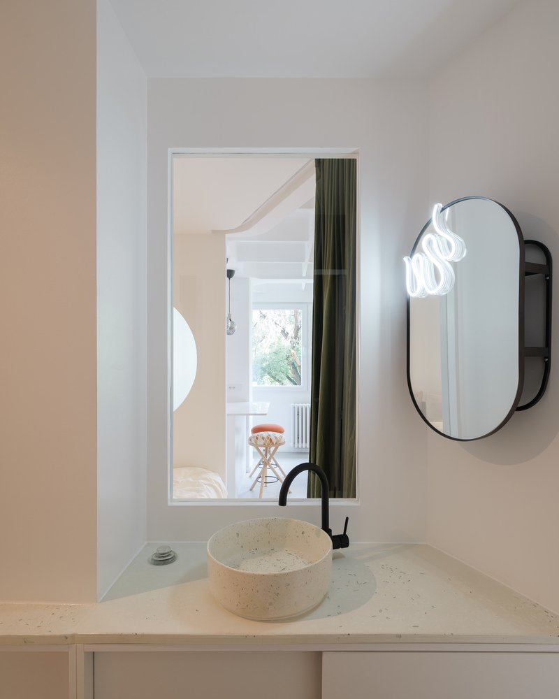 Bathroom: terrazzo basin on a white counter, rectangular and oval mirror composition, view back into the living room through the circular window reveal