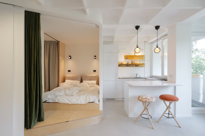 Wide view: kitchen with three pendant lights at right, bedroom platform raised above the main floor, green velvet curtain drawn back, white coffered ceiling