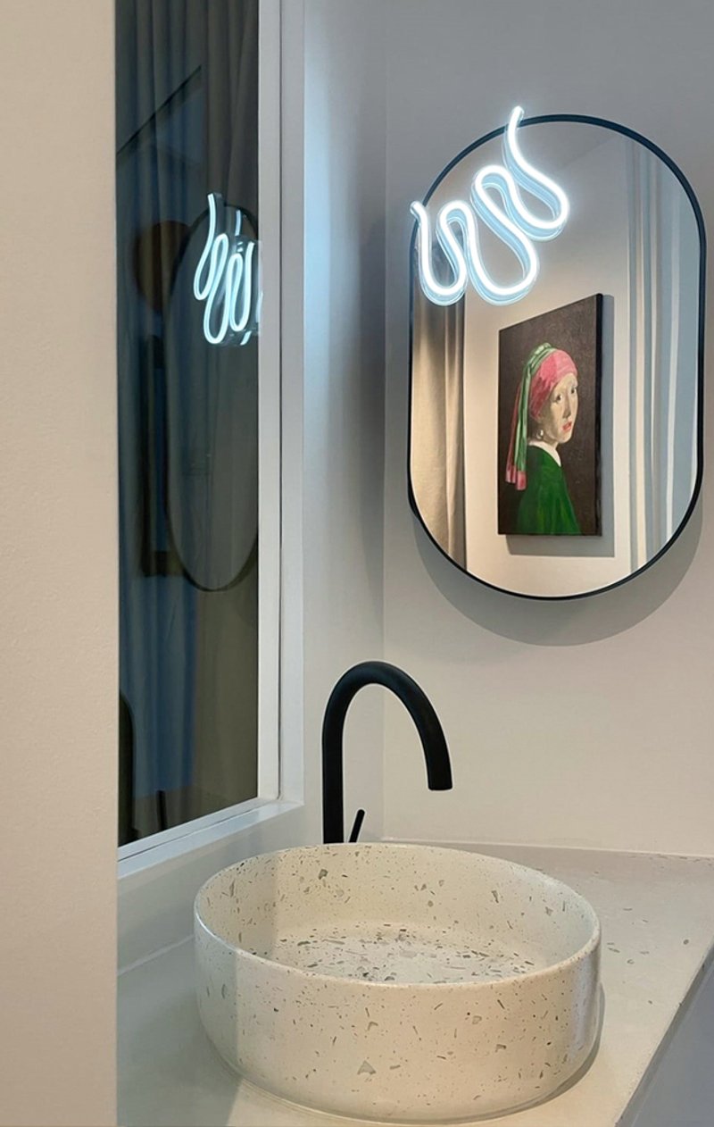 Bathroom accent: neon cursive sign reflected in the oval mirror, framed portrait in a pink frame, terrazzo basin below
