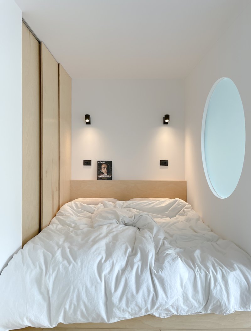 Bedroom close-up: white bedding and pillows, two matching black wall lamps, timber headboard, round window on the right wall