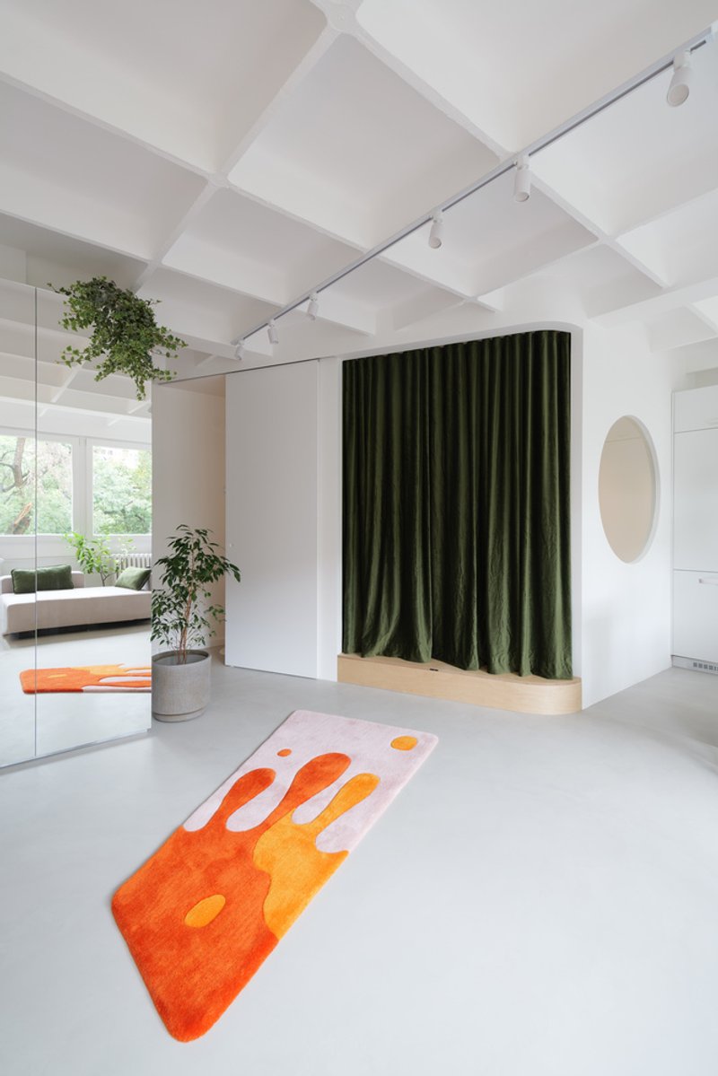 Living room with rug: orange-on-pink kite-shaped rug on the polished white floor, green velvet curtain behind, round pink mirror, plants cascading from above