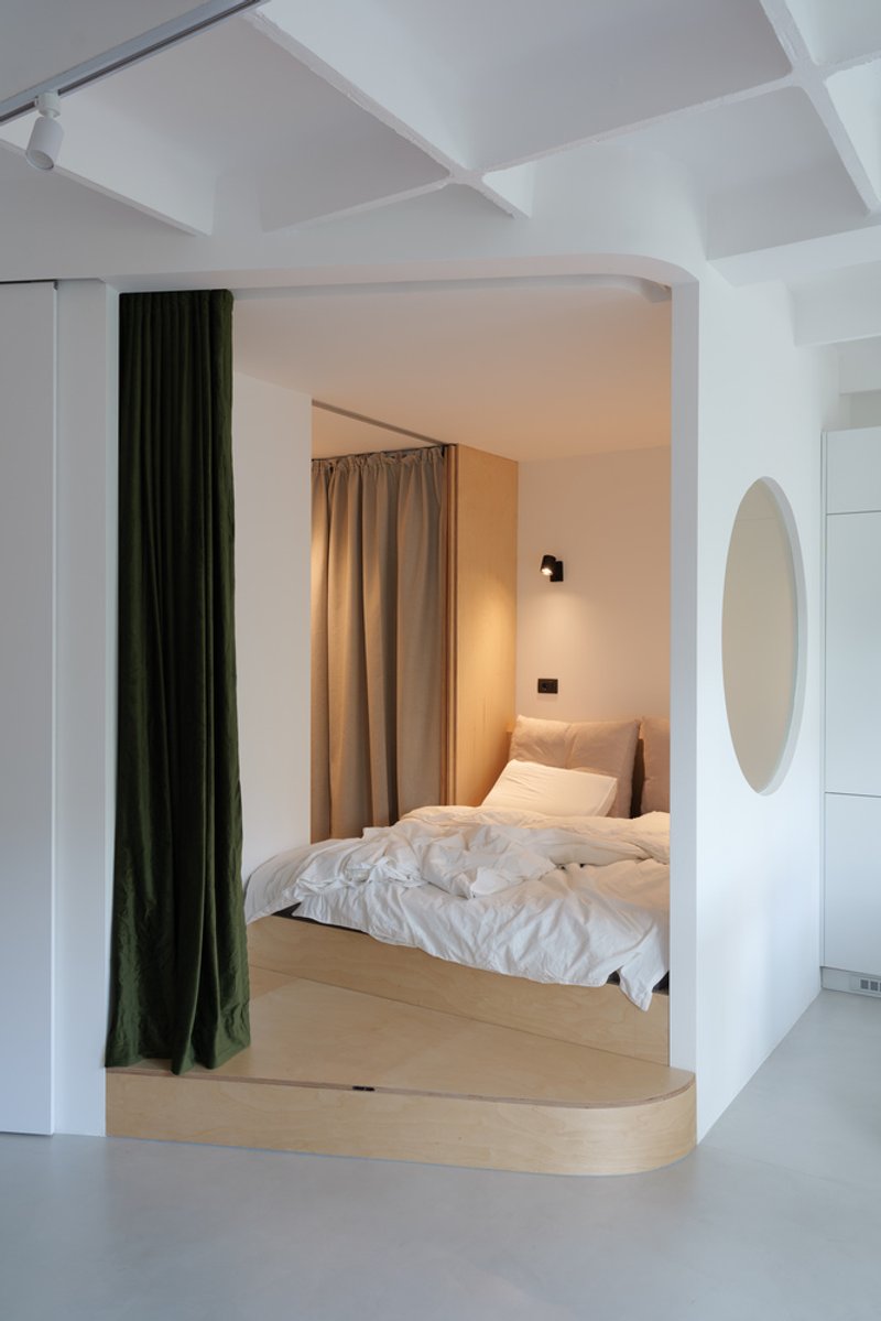 Embedded bedroom: raised timber platform with a white bed, green velvet curtain as a movable partition, round mirror on the wall, wall sconce