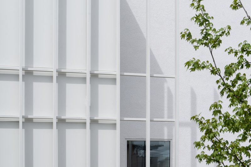 Facade close-up: white metal fin rhythm, small window set deep in the wall, tree shadow on the surface