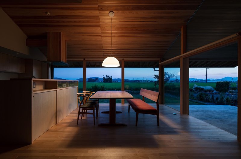 Dining at dusk: cedar interior glowing under pendant lamp, mountains silhouetted through full-width window, deep blue sky