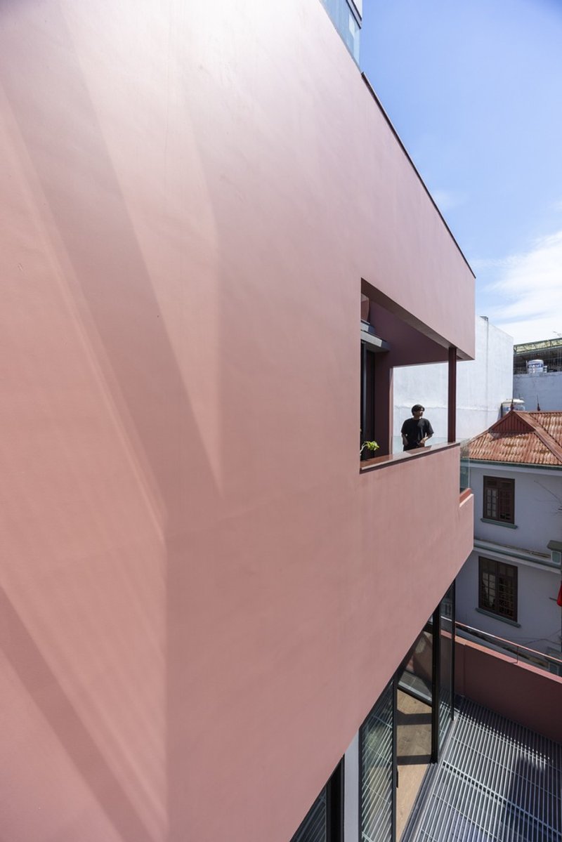 Pink rendered facade with recessed window where a figure stands overlooking neighboring rooftops