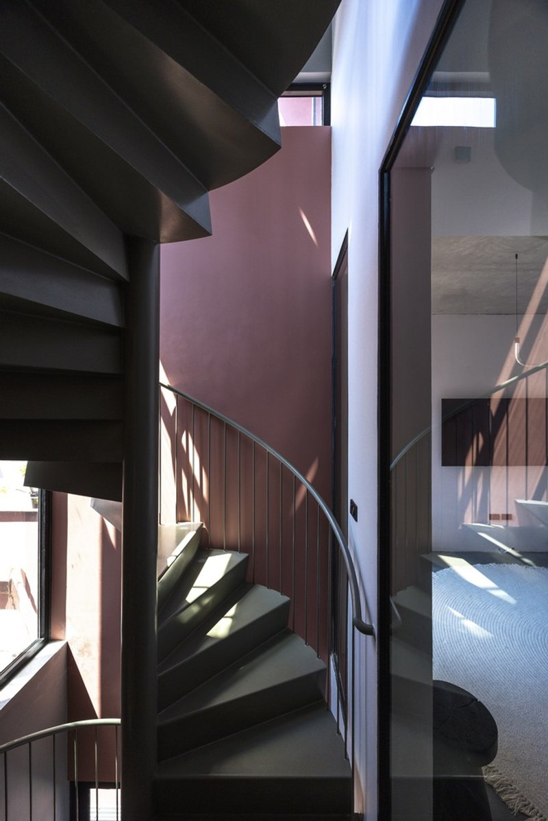 Steel spiral staircase with timber paneling ascending through a double-height space with filtered afternoon light