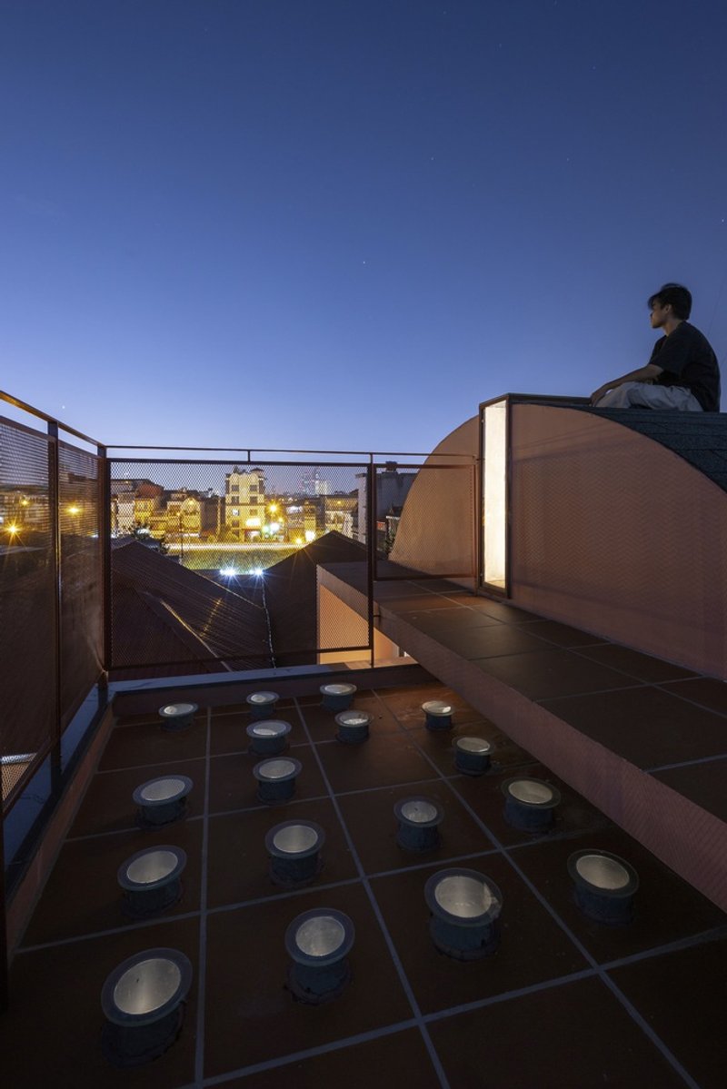 Rooftop terrace at twilight with person seated on curved illuminated volume and skylights set into tile floor