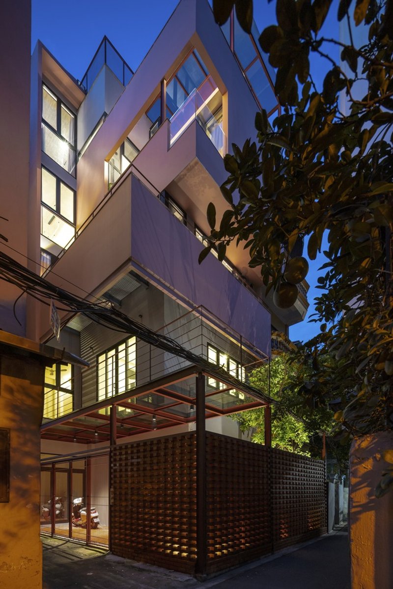 Street facade at dusk showing cantilevered volumes above perforated brick screen and entry canopy beside leafy branches