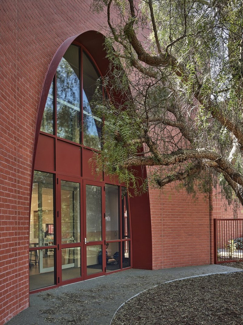 Arched entrance with glazed metal doors set into red brick facade beneath overhanging tree limbs