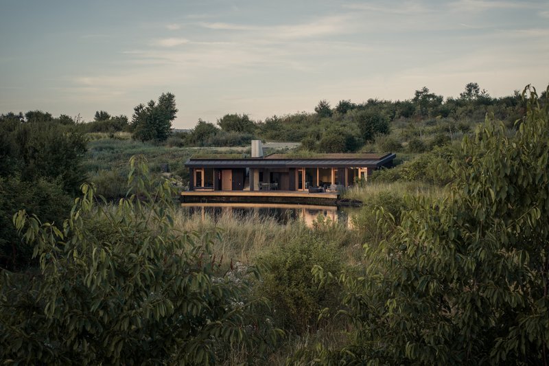 From the meadow: low dark house with timber deck, wildflower planting in foreground, trees, flat Hungarian plain, evening sky