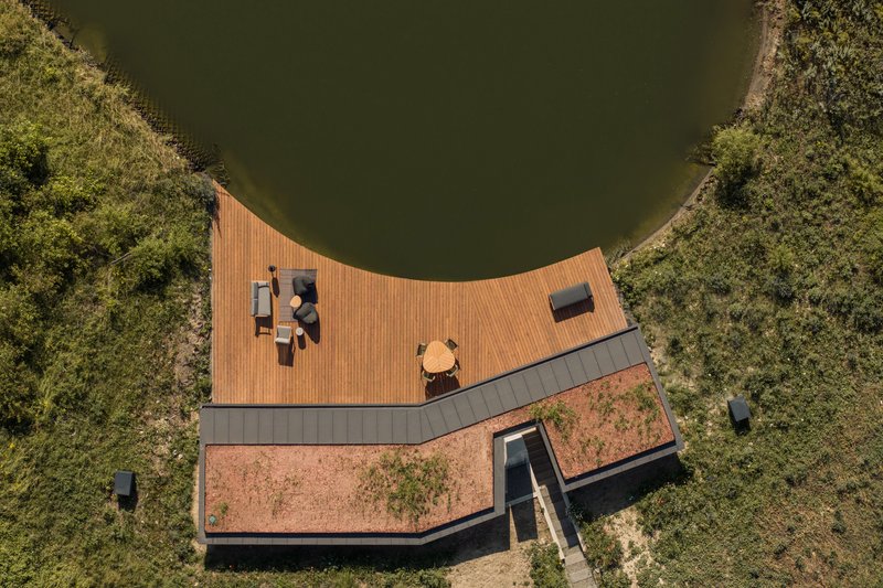 Aerial: V-shaped plan with timber deck wrapping the pond edge, green roof, concrete chimney, meadow surrounding