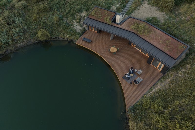 Aerial: V-shaped house with timber deck curving along the pond edge, green roof, two figures sitting on the deck, meadow