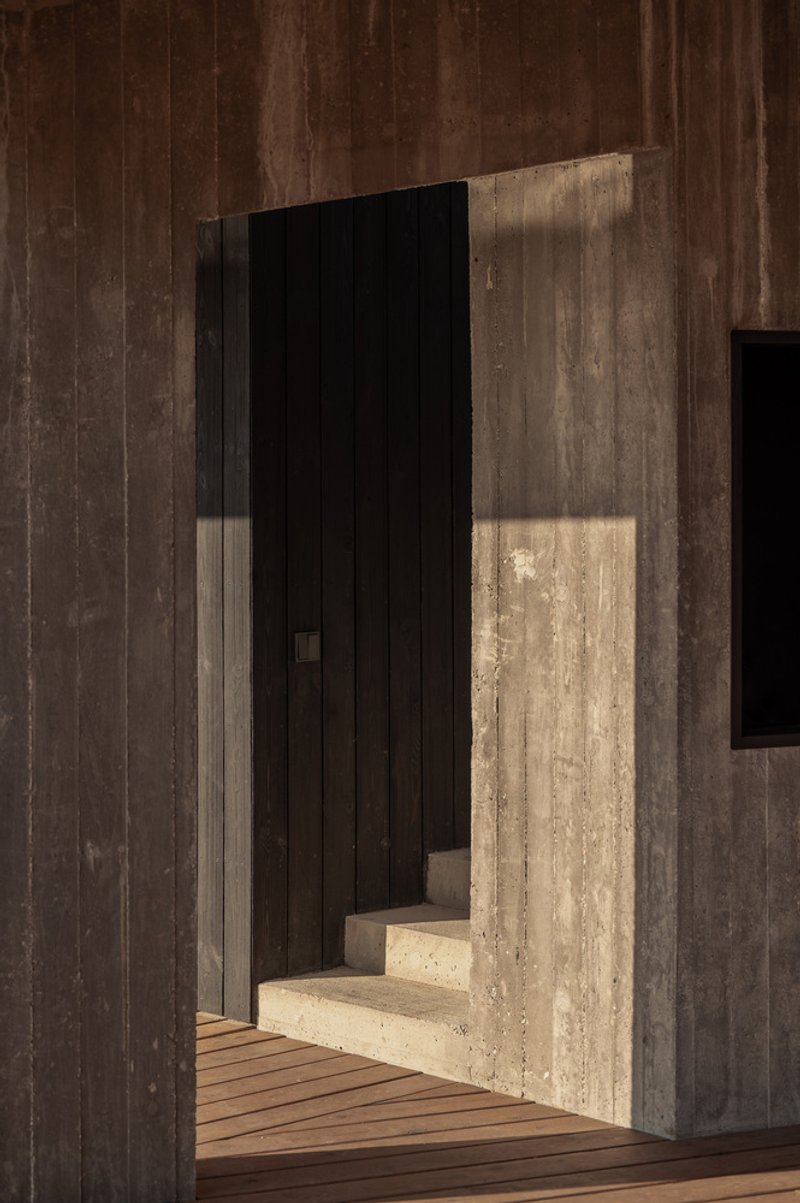 Entry passage: charred timber walls on both sides, concrete step, sharp shadows, timber deck ahead