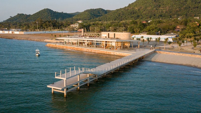 Timber walkway stretching from shore to the glass-walled pavilion at golden hour with forested hills beyond