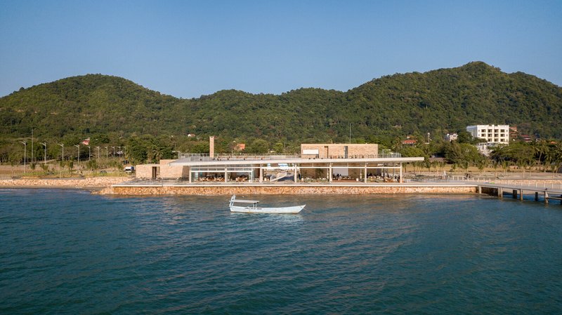 Waterfront view of the low-slung pavilion with exposed concrete columns and brick retaining wall under forested hills