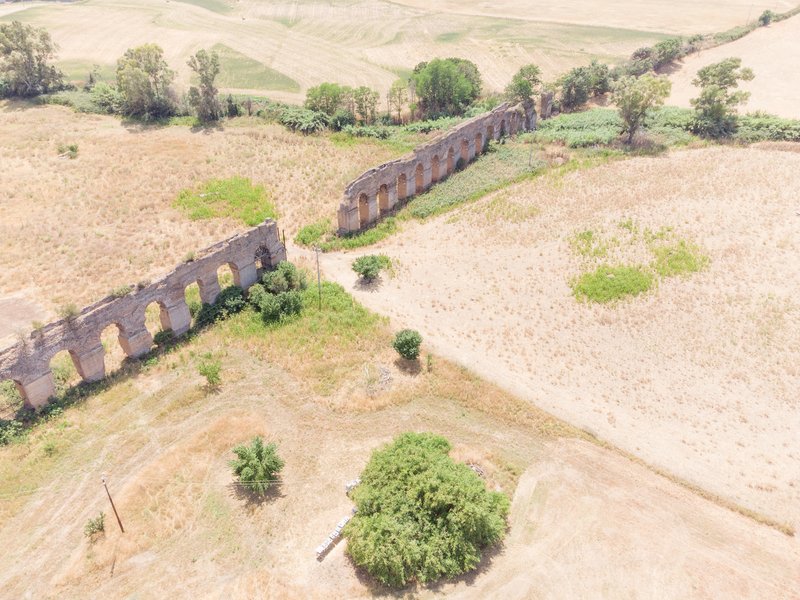 Aerial view of two ruined brick aqueduct walls cutting through cultivated farmland with scattered trees