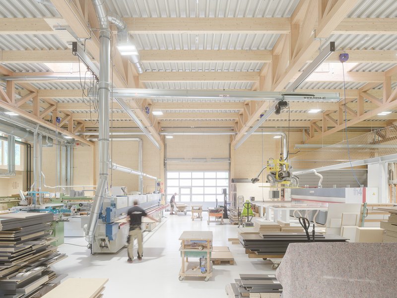 Workshop interior with exposed timber beams, metal ducts, work tables and a person walking through sunlight
