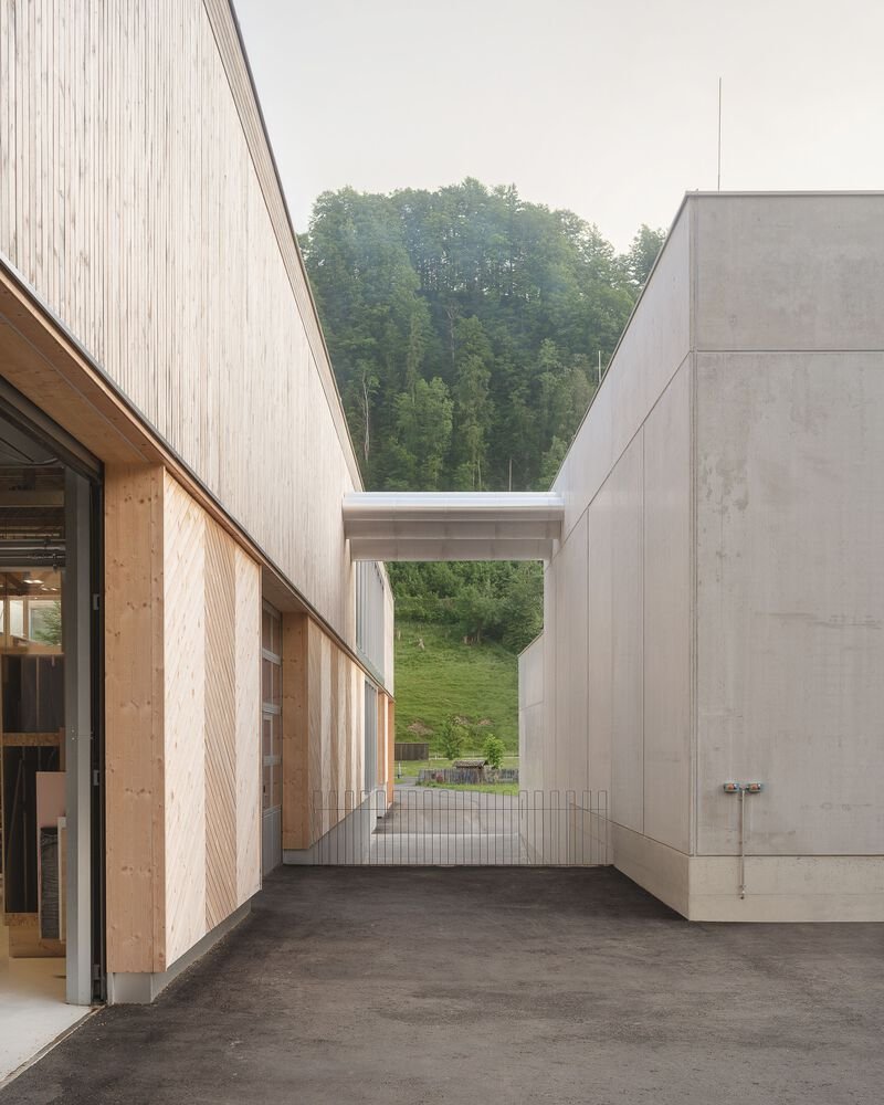 Exterior passageway between timber-clad workshop and concrete volumes framing a view toward forested hills