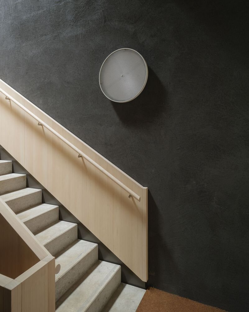 Concrete staircase with pale timber handrail against a dark wall with circular mesh speaker