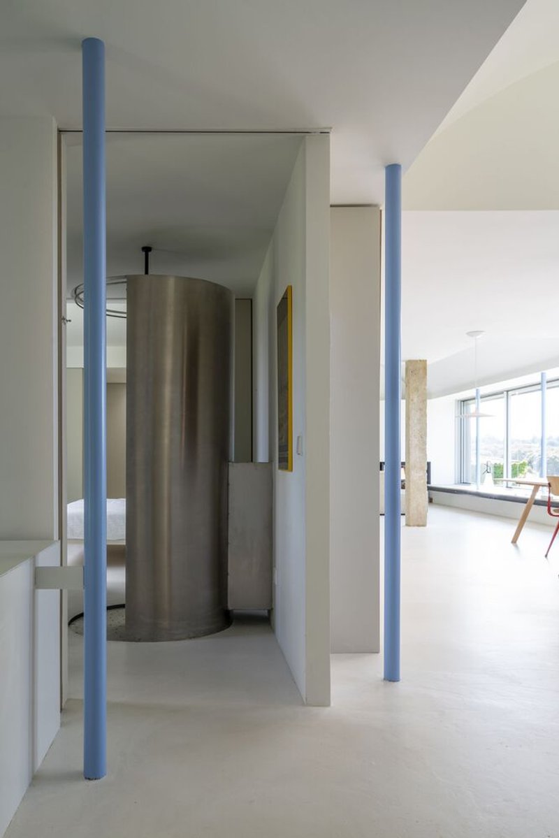 White interior with blue cylindrical columns framing a view of a freestanding curved metal shower enclosure
