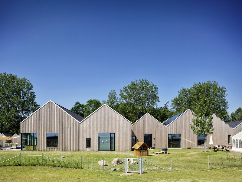 Row of timber-clad gable-roofed volumes fronting a grass lawn with playground equipment and young trees