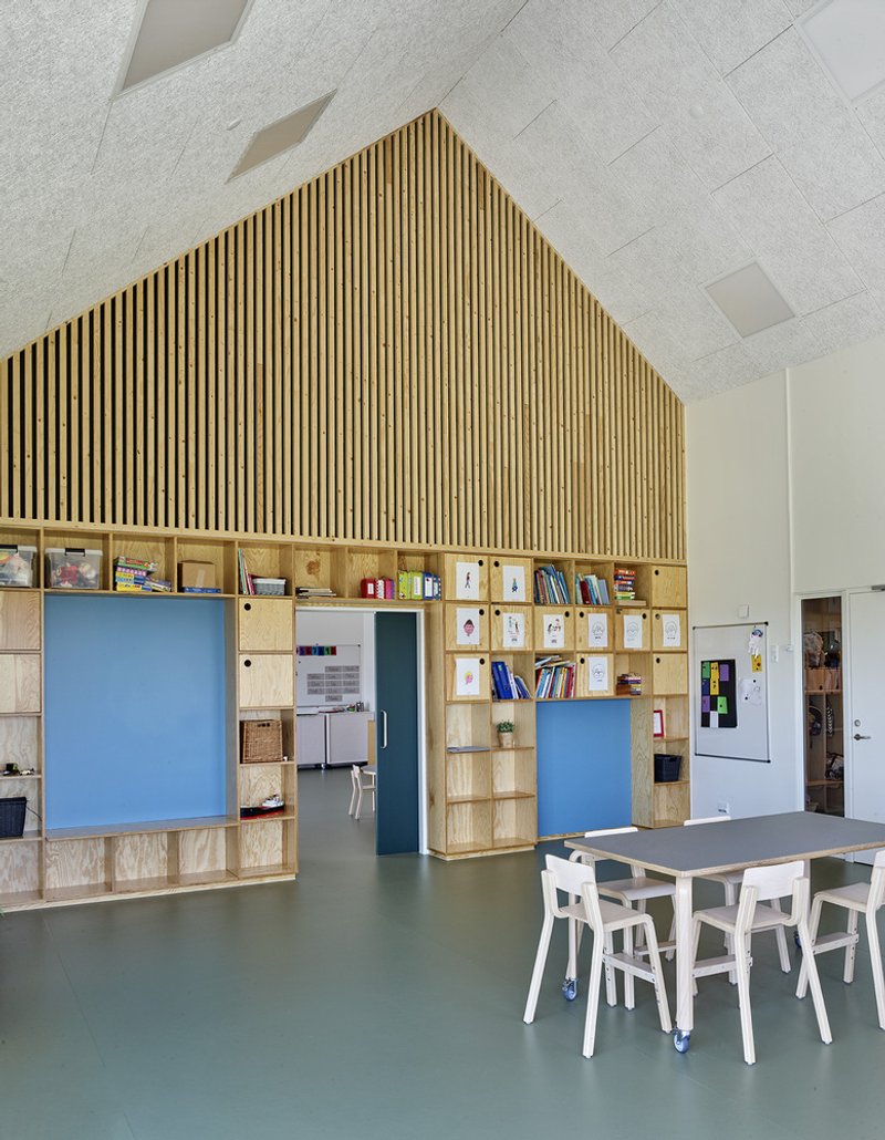 Classroom space with house-shaped storage unit featuring slatted timber gable and built-in plywood cubbies with sliding panels