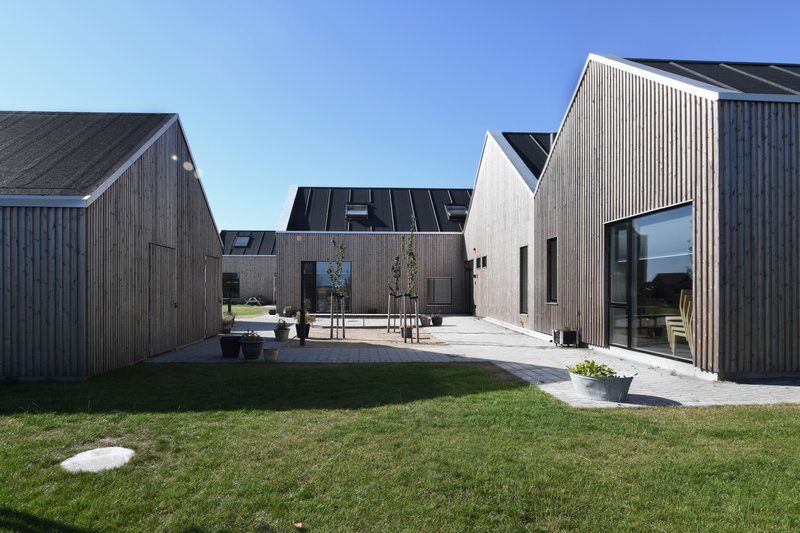 Cluster of vertically clad timber volumes arranged around a shared lawn and paved courtyard under clear skies