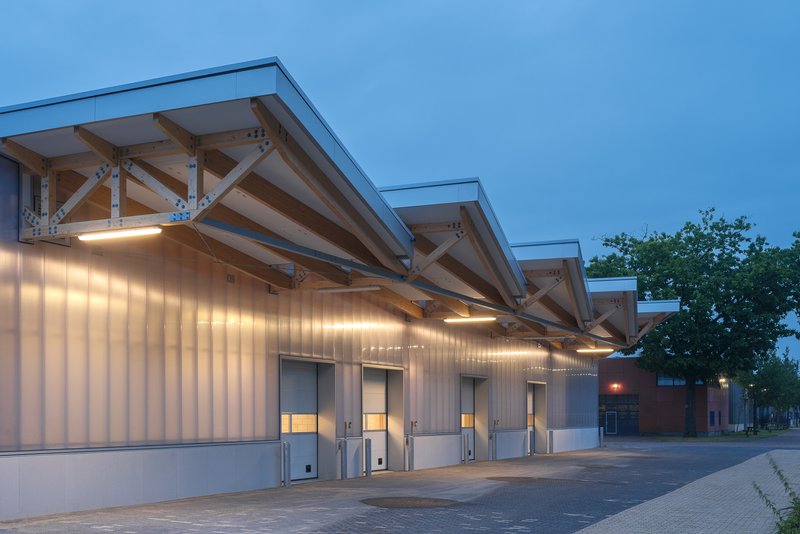 Repeating sawtooth roofline with exposed timber trusses above a ribbed metal facade at twilight