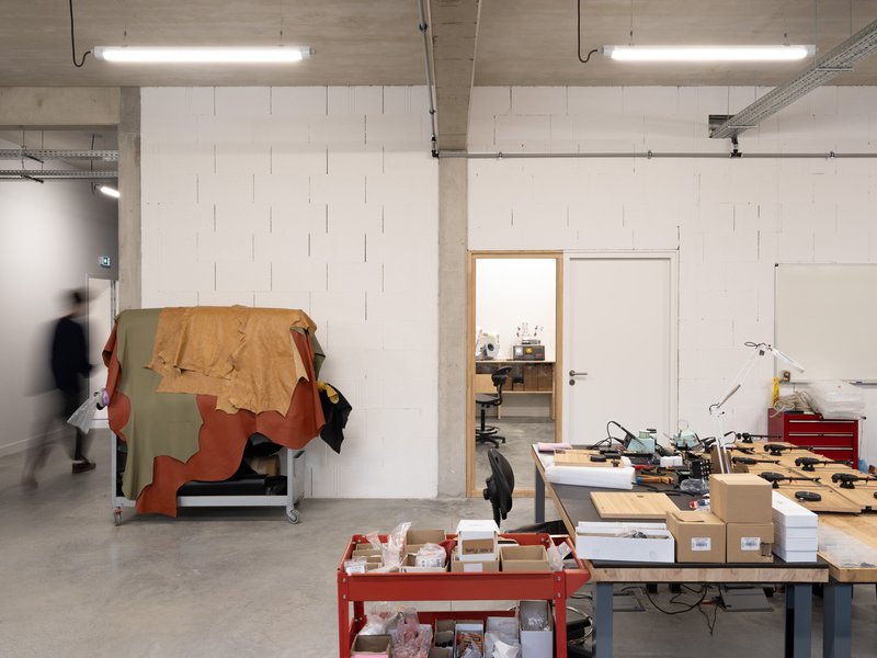 Workshop interior with exposed concrete ceiling, fluorescent tubes, and a person moving past worktables and stored materials