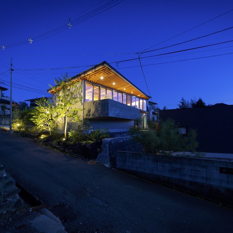 Street view at night: the house glowing warm on its slight rise above the road, timber rafter ceiling visible through the full-height glazing, power lines and trees in the foreground