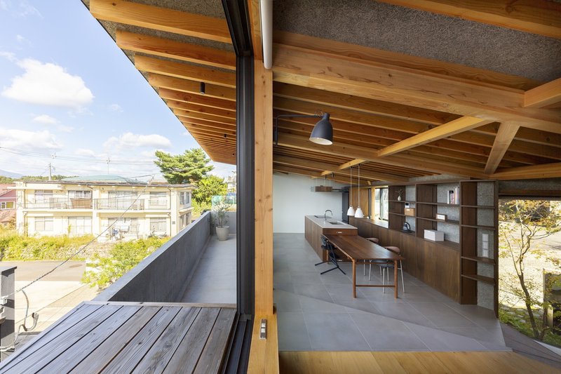 Upper living room: exposed timber rafter ceiling, long dining table, concrete floor, dark timber storage wall, full-height glass opening to the balcony with distant hills