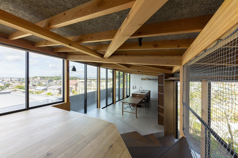 Upper open plan: dense timber rafter ceiling, dining table, concrete floor, wire mesh stair balustrade at right, full-width glazing framing the town view