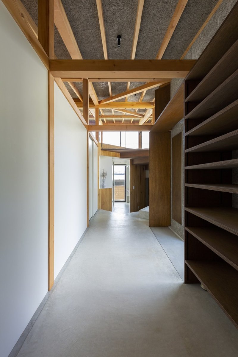 Corridor: timber shelving on the right, exposed rafter ceiling above, concrete floor, view through to a brighter room at the end