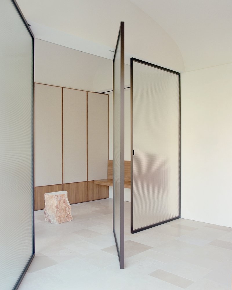 Pivoting translucent glass door framed in dark metal between rooms with pale stone floors