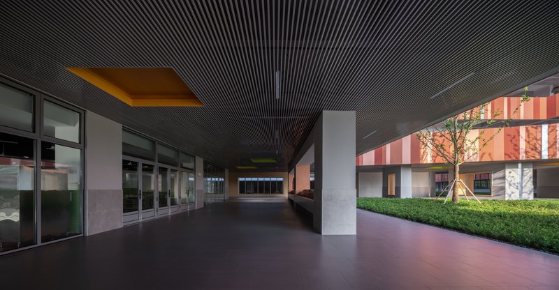 Covered corridor with dark slatted ceiling and square columns leading toward illuminated courtyard facades at dusk