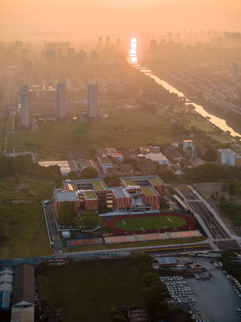 Aerial view of the school campus with vertical striped cladding and green rooftop sports field at golden hour