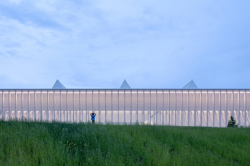 Perforated metal facade with triangular roofline and figure in tall grass at dusk