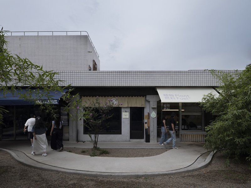 Street-level facade with white mosaic tile upper volume and retail storefronts beneath shallow canopy at dusk