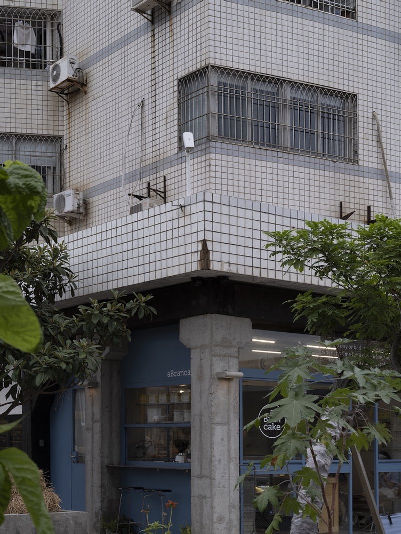 Corner shopfront with exposed concrete columns and blue-grey facade set below white mosaic tile residential structure