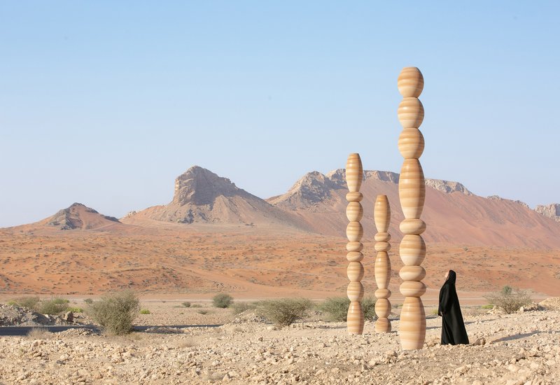 Three stacked timber columns in a desert landscape with a figure in black and distant rocky mountains