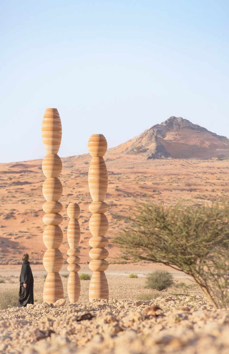 Three laminated wood columns beside a lone tree with a figure at the base and mountain beyond