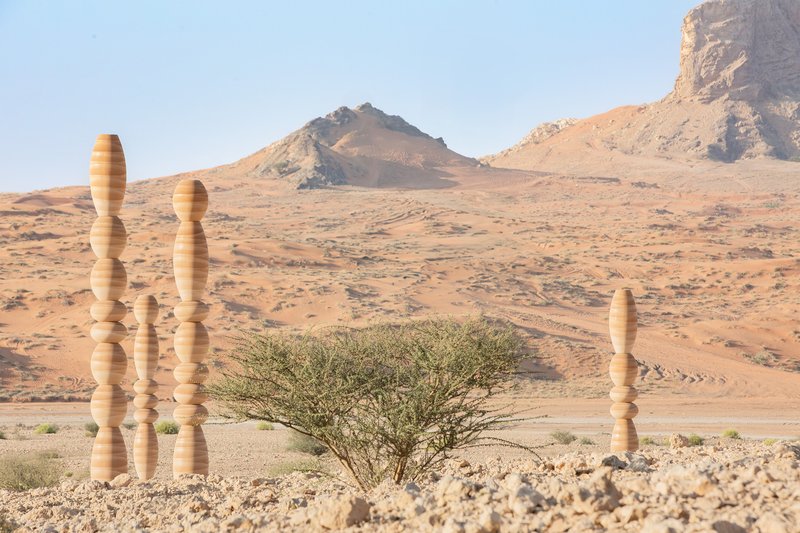 Stacked timber columns with spherical joints standing beside low desert shrubs at the base of rocky hills